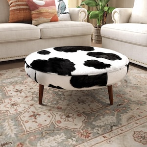 May include: A round ottoman featuring a black and white cow print pattern on its upholstered top and sides. The piece is supported by four tapered wooden legs in a medium brown finish, providing a stable base for this circular furniture item.