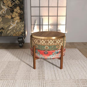 May include: A round ottoman featuring a dark wood top with a gold metal rim, supported by four brown wooden legs. The sides are upholstered in a multi-colored fabric with intricate patterns in shades of green, red, orange, and cream.