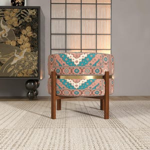 a colorful, patterned chair with a wooden frame, placed on a beige rug in a room with a large window and a decorative cabinet.
