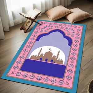 May include: A prayer rug with a pink border and a blue and pink floral design. The center features a silhouette of a mosque against a light purple background. The rug is rectangular and has a blue border.