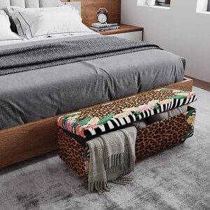 May include: A rectangular storage bench with a leopard print design on the sides and top. The top features a floral and zebra stripe pattern. The bench is open, revealing folded blankets and a gray scarf. The bench is placed at the foot of a bed.