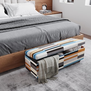 May include: A rectangular storage ottoman with a striped pattern in shades of blue, orange, gray, and white sits at the foot of a bed. A gray blanket and pillows are visible on the bed. A gray scarf hangs from the ottoman.
