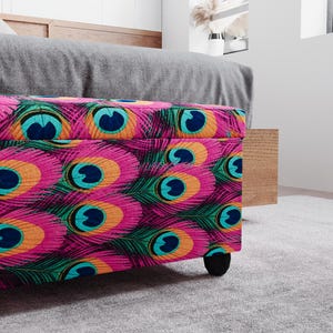 a colorful, patterned ottoman or storage box with a vibrant design of peacock feathers in shades of pink, blue, green, and orange. The ottoman is placed on a gray carpeted floor, and there is a wooden cabinet visible in the background.
