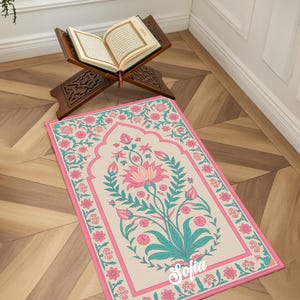 May include: A prayer rug with a floral design in pink, teal, and cream colors. The rug features a central floral motif and a pink border. The word "Sofia" is written on the rug. An open book rests on a wooden stand in the background.