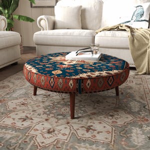 May include: A round ottoman featuring a deep blue top with a multi-colored geometric pattern and a rust-orange side panel with a repeating diamond motif. The piece is supported by four tapered dark brown wooden legs and holds a white book.