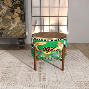 Puede incluir: Un otomán redondo con tapa y patas de madera. La tela presenta un fondo verde con estampados florales, de leopardo y geométricos. La tapa de madera es de color marrón claro. El otomán mide unos 40 cm de diámetro.