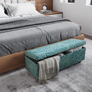 May include: A teal and white patterned storage bench is positioned at the foot of a bed with gray bedding. The bench is open, revealing folded blankets and a gray scarf. The bed has a wooden frame.