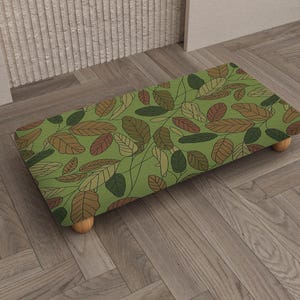 Pode incluir: Um pufe retangular com fundo verde e um padrão de folhas em tons de castanho, verde e bege. O pufe tem quatro pequenas pernas de madeira arredondadas. O design lembra uma impressão botânica.