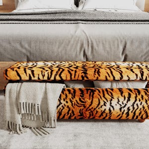May include: A rectangular storage bench featuring a vibrant orange and black tiger stripe pattern. The lid is partially lifted, revealing white fabric items stored inside. A light gray fringed throw blanket hangs over the left side of the bench.