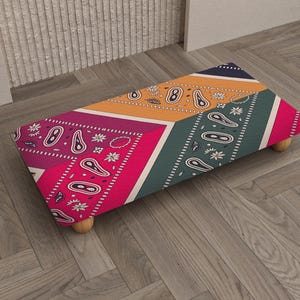 Könnte beinhalten: Ein rechteckiger Hocker mit einem farbenfrohen Patchwork-Design. Die Oberseite zeigt eine Mischung aus rosa, orangefarbenen und türkisfarbenen Paisley-Mustern. Der Hocker wird von vier kleinen, abgerundeten Holzbeinen getragen. Das Design ist lebendig und auffällig.