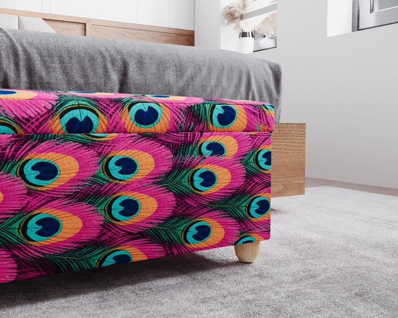 a colorful, patterned ottoman or storage box with a vibrant design of peacock feathers in shades of pink, blue, green, and orange. The ottoman is placed on a gray carpeted floor, and there is a wooden cabinet visible in the background.
