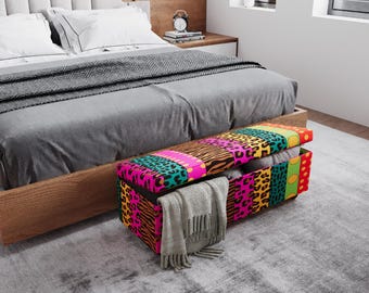 May include: A colorful storage bench with a patterned design sits at the foot of a bed. The bench features a variety of animal prints and bright colors. A gray blanket and other items are stored inside. The bed has a gray comforter and white pillows.