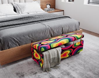 May include: A colorful storage ottoman with a psychedelic pattern of swirling shapes in red, yellow, green, and purple. The ottoman is open, revealing folded blankets and a gray scarf draped over the edge. It sits at the foot of a bed.