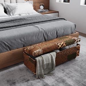 Puede incluir: Una cama con edredón y almohadas grises, una manta estampada y un banco de almacenamiento al pie. El banco tiene un diseño de patchwork con estampados de animales y está abierto, revelando mantas dobladas y una bufanda gris.