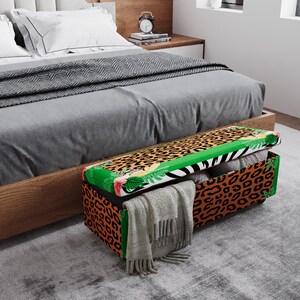 May include: A decorative storage bench with a leopard print exterior and a green border. The lid features a zebra stripe and tropical floral design. A gray blanket is draped over the front of the bench, which is placed at the foot of a bed.