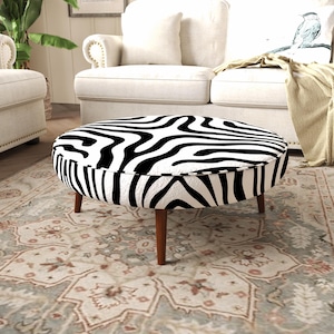 May include: A round ottoman featuring a black and white zebra stripe pattern on the top and sides. The piece is supported by four tapered wooden legs in a medium brown finish, providing a stable base for this circular furniture item.