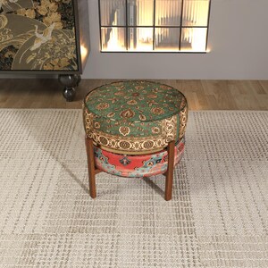 a round, colorful ottoman with a patterned design, placed on a white rug in a room with a bookshelf and a window.