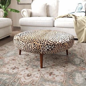 Op de afbeelding: Een ronde poef met een luipaardprint in tinten beige, zwart en bruin. Het meubelstuk wordt ondersteund door vier taps toelopende houten poten in een middenbruine afwerking, wat een stabiele basis vormt voor het ronde, gestoffeerde bovenvlak.