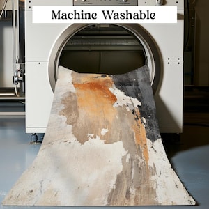May include: A white washing machine with the words "Machine Washable" on the front. A large, abstract patterned rug is being fed into the machine.