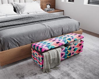 May include: A colorful storage bench with a geometric pattern in pink, blue, and yellow sits at the foot of a bed. The bench is open, revealing folded blankets and a gray scarf. The bed has a gray comforter and white pillows.