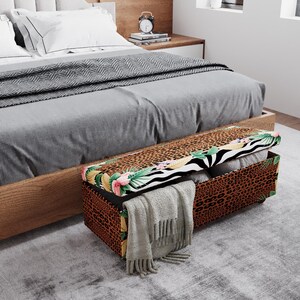 May include: A decorative storage bench with a leopard print exterior and a tropical floral and zebra print on the lid. The bench is open, revealing folded blankets and a gray scarf. The bench is placed at the foot of a bed.