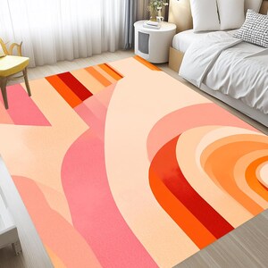 May include: Abstract area rug with a geometric design in shades of orange, pink, and cream. The rug features a large, curved shape with concentric circles in shades of orange.