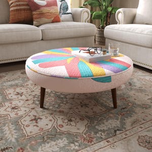 May include: A round ottoman with a light pink base and four dark brown wooden legs. The top surface features a quilted fabric pattern with radiating segments in bright colors including yellow, pink, blue, and purple. A book and glasses sit on top.