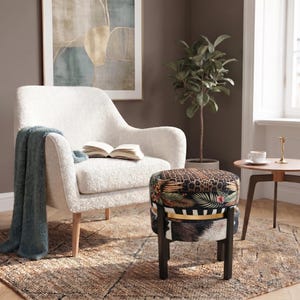 Op de afbeelding: Een crèmekleurige fauteuil met een textuurrijke stoffen afwerking, voorzien van een blauwe plaid en een open boek op de zitting. Daarnaast staat een ronde poef met een veelkleurig patroon, een gouden band en donkerhouten poten.