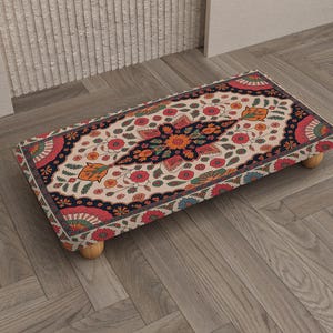 May include: A rectangular footstool with a floral pattern in red, orange, and blue on a cream background. The footstool has a dark navy border and is supported by four small wooden legs. The design is reminiscent of a traditional rug.
