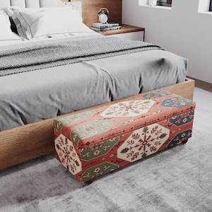 May include: A patterned storage bench in red, green, and blue sits at the foot of a bed. The bed has a light wood frame, grey bedding, and white pillows. A grey rug covers the floor. The bench is approximately 120 cm long.
