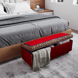 May include: A red storage bench with a leopard print trim and a hinged lid. The bench is open, revealing folded blankets and a gray scarf. The bench is placed at the foot of a bed with gray bedding.
