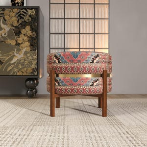 a vibrant, patterned chair with a wooden frame, placed on a white rug in a room with a large window and a decorative cabinet.