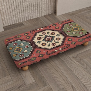 Könnte beinhalten: Ein rechteckiger Fußhocker mit einem gemusterten Design in Rot, Türkis, Grün und Creme. Der Fußhocker hat einen Holzrahmen und ruht auf vier kleinen, abgerundeten Holzbeinen. Das Muster zeigt geometrische Formen und florale Motive.