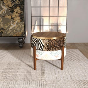 Op de afbeelding: Een ronde poef met een donkerhouten blad en een goudkleurige metalen rand. Het bovenste gedeelte is bekleed met een zwart-witte zebraprint en een bruin-zwarte luipaardprint. Het onderste gedeelte heeft een witte stof met een kleurrijk bloemenpatroon.