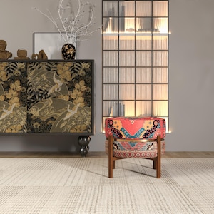 a modern living room with a vibrant red and blue patterned chair, a black and gold cabinet with a floral design, and a glass-paneled wall with a white vase and branches.