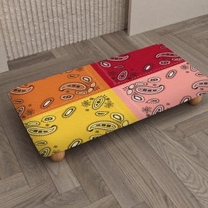 Könnte beinhalten: Ein rechteckiger Hocker mit einem farbenfrohen Paisley-Muster. Die Oberseite ist in vier Abschnitte unterteilt: Orange, Gelb, Rot und Rosa. Jeder Abschnitt weist ein weißes Paisley-Design auf. Der Hocker hat vier kleine, runde Holzbeine.