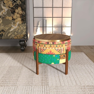 Puede incluir: Un taburete redondo con tapa de madera oscura y borde de metal dorado. La base está tapizada en tela multicolor con patrones geométricos en amarillo, rojo y verde, con detalles de estampado de leopardo, sobre cuatro patas de madera.