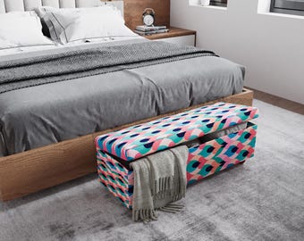 May include: A colorful storage bench with a geometric pattern in pink, blue, and teal sits at the foot of a bed with gray bedding. A gray blanket is draped over the open bench, revealing storage space. The bed has a wooden frame.