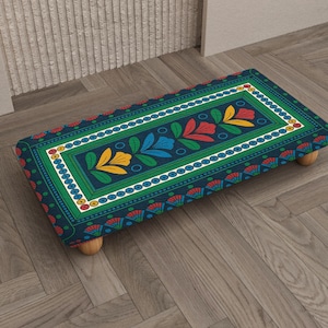 May include: A rectangular footstool with a navy blue background and a colorful floral design. The design includes yellow, red, and blue flowers, with a white border. The footstool has four wooden legs and is placed on a wooden floor.