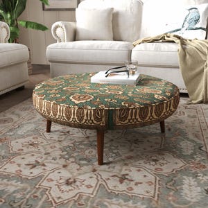 May include: A round ottoman with a green and brown floral pattern on the top and a brown paisley border along the side. The piece features four tapered wooden legs and supports a white book with black glasses and a clear glass on top.