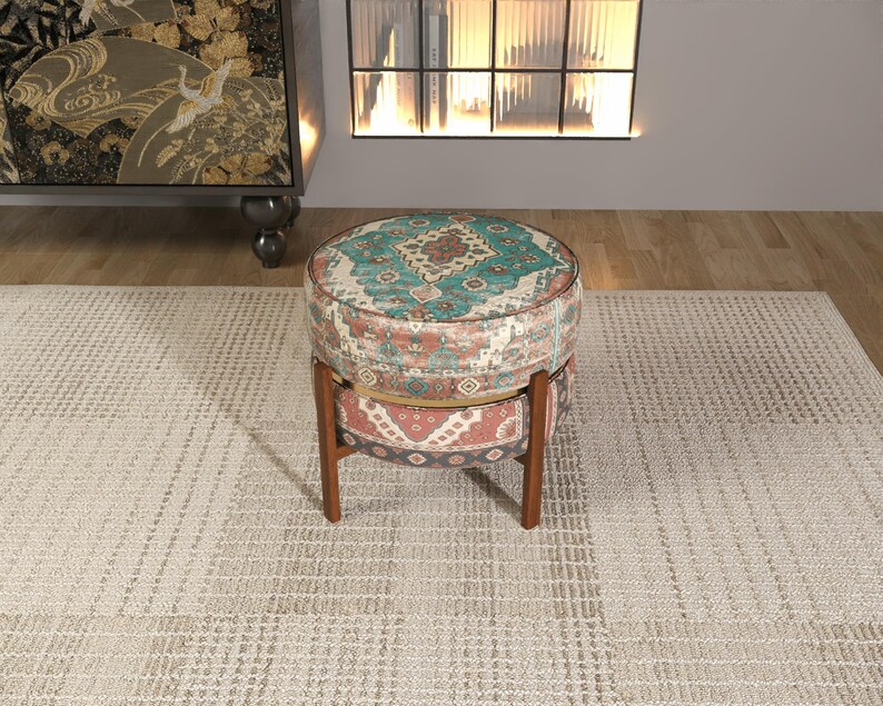 a round, colorful ottoman with a patterned design, placed on a beige rug in a room with a fireplace and a bookshelf.