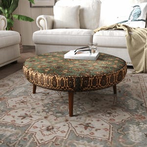 Può includere: Un pouf rotondo caratterizzato da una parte superiore verde con un motivo floreale scuro e un pannello laterale con un design paisley marrone e oro. Il mobile poggia su quattro gambe in legno affusolate e sostiene un libro bianco con occhiali.