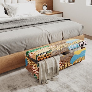 May include: A rectangular storage bench featuring a vibrant patchwork of animal prints, geometric shapes, and wavy blue patterns. The lid is partially open, revealing a white interior, and a beige fringed throw blanket hangs over the front side.