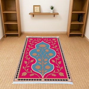 May include: A rectangular prayer rug with a vibrant pink background and a central design in teal and tan. The design features a stylized floral motif. The rug has a black border with tan and blue fringe.