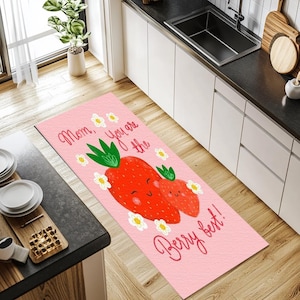 Puede incluir: Una alfombra de cocina rosa con un diseño de fresas y el texto "Mom, You are the Berry best!". La alfombra presenta dos fresas sonrientes, hojas verdes y flores blancas.