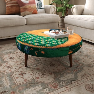 May include: A round ottoman featuring a vibrant peacock feather pattern in shades of green, blue, and gold on the top surface. The sides are dark green with floral accents, supported by four tapered wooden legs. A book and glasses rest on top.