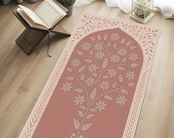 Op de afbeelding: Een gebedskleed met een bloemenboomontwerp in roze en beige tinten. Het kleed heeft een gewelfde bovenkant en een rand met bladerpatronen. Het ontwerp is gecentreerd op een roze achtergrond.