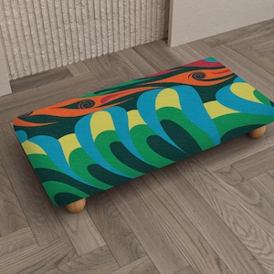 May include: A rectangular footstool with a colorful, abstract pattern. The design features swirling shapes in shades of green, blue, yellow, and orange against a dark background. The stool has four wooden legs and is placed on a wooden floor.