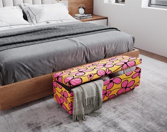 May include: A yellow storage bench with a pink floral pattern sits at the foot of a bed. The bench is open, revealing blankets and a gray scarf. The bed has a gray comforter and white pillows.
