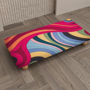 May include: A rectangular footstool with a vibrant, retro-style design. The top features a swirling pattern of red, orange, yellow, blue, and green. It has four short, rounded wooden legs. The footstool is placed on a wooden floor.